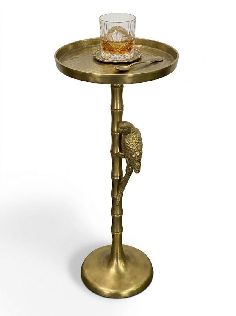 May include: A gold-colored side table with a round top and a parrot perched on a bamboo-style stem. A glass of amber liquid sits on the table, along with a small coaster and spoon. The table has a hammered texture.