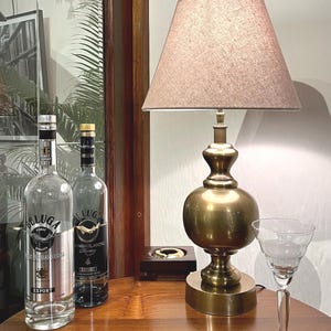 Könnte beinhalten: Eine Messing-Tischlampe mit einem strukturierten beigen Lampenschirm, zwei klaren Beluga-Wodkaflaschen aus Glas mit silbernen und schwarzen Etiketten sowie einem klaren Stielglas. Die Lampe hat einen runden Fuß und einen bauchigen Schaft.