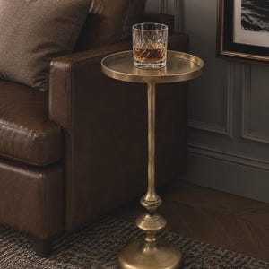 Op de afbeelding: Kleine, antieke messing bijzettafel met een rond blad en een enkele stam. Een geslepen kristallen glas met amberkleurige vloeistof staat op de tafel. De tafel staat naast een bruine leren bank.
