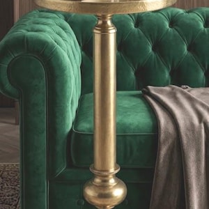Puede incluir: Una mesa auxiliar dorada con una tapa redonda sostiene un vaso de líquido ámbar, un libro y un teléfono. La mesa está junto a un sofá de terciopelo verde esmeralda con mechones. Una manta marrón cubre el sofá.