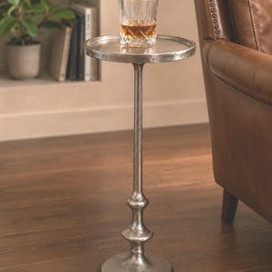 May include: A silver-toned side table with a round top and a tall, slender stem. A glass of amber liquid sits on the table. The table has a decorative base and is placed on a wooden floor.