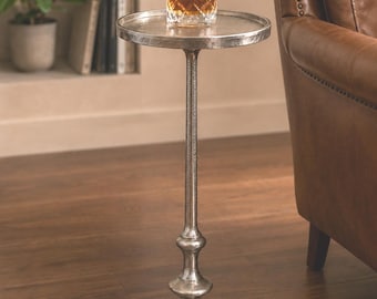 Può includere: Un tavolino argentato con piano rotondo e stelo alto. Un bicchiere di liquido ambrato è appoggiato sul tavolo. Il tavolo ha una base decorativa ed è posto su un pavimento in legno. Altezza circa 76 cm.