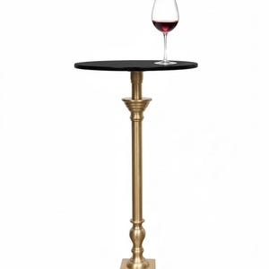 May include: A gold-colored, pedestal table with a black circular top. A glass of red wine sits on the table. The table has a square base and a long, ornate stem.