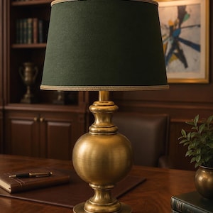 May include: A table lamp featuring a brushed brass base with a rounded, bulbous center and a circular pedestal. The lamp is topped with a dark forest green fabric drum shade, which is finished with a gold-toned braided trim along the edges.