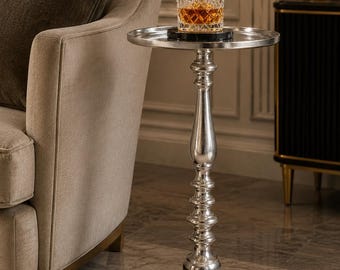Può includere: Un tavolino argentato con piano rotondo e stelo ornato. Un bicchiere di liquido ambrato poggia su un sottobicchiere nero sul tavolo. Il tavolo è accanto a una poltrona beige. Il tavolo è alto circa 70 cm.