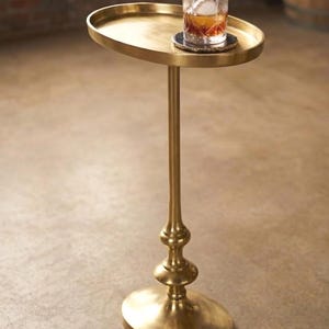 Puede incluir: Una mesa auxiliar dorada con tapa y base ovaladas. La mesa tiene un tallo delgado y ornamentado. Un vaso de líquido ámbar con hielo descansa sobre un posavasos. La mesa mide aproximadamente 71 cm de altura.