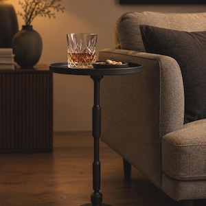 May include: A tall, slender black metal side table with a circular top and a weighted base. The table features a central column with decorative cylindrical accents. A clear, faceted glass tumbler containing an amber liquid sits on the surface.