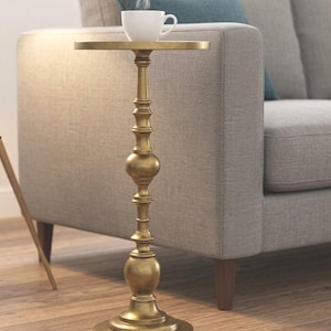 Puede incluir: Una mesa auxiliar dorada con una parte superior redonda y un tallo ornamentado y detallado. Una taza de té blanca con vapor está sobre la mesa. La mesa está junto a un sofá gris con un cojín azul. La mesa mide aproximadamente 61 cm de altura.
