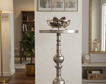 Può includere: Un alto tavolo a piedistallo color argento con piano rotondo e base decorativa a più livelli. Una ciotola di vetro riempita con pietre grigie lisce e due pigne marroni è posizionata al centro della superficie circolare del tavolo.
