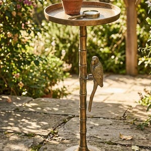 May include: A bronze-colored plant stand with a round top, a bamboo-style stem, and a parrot figurine. A small potted plant and a magnifying glass rest on the top. The stand is outdoors on a stone patio.