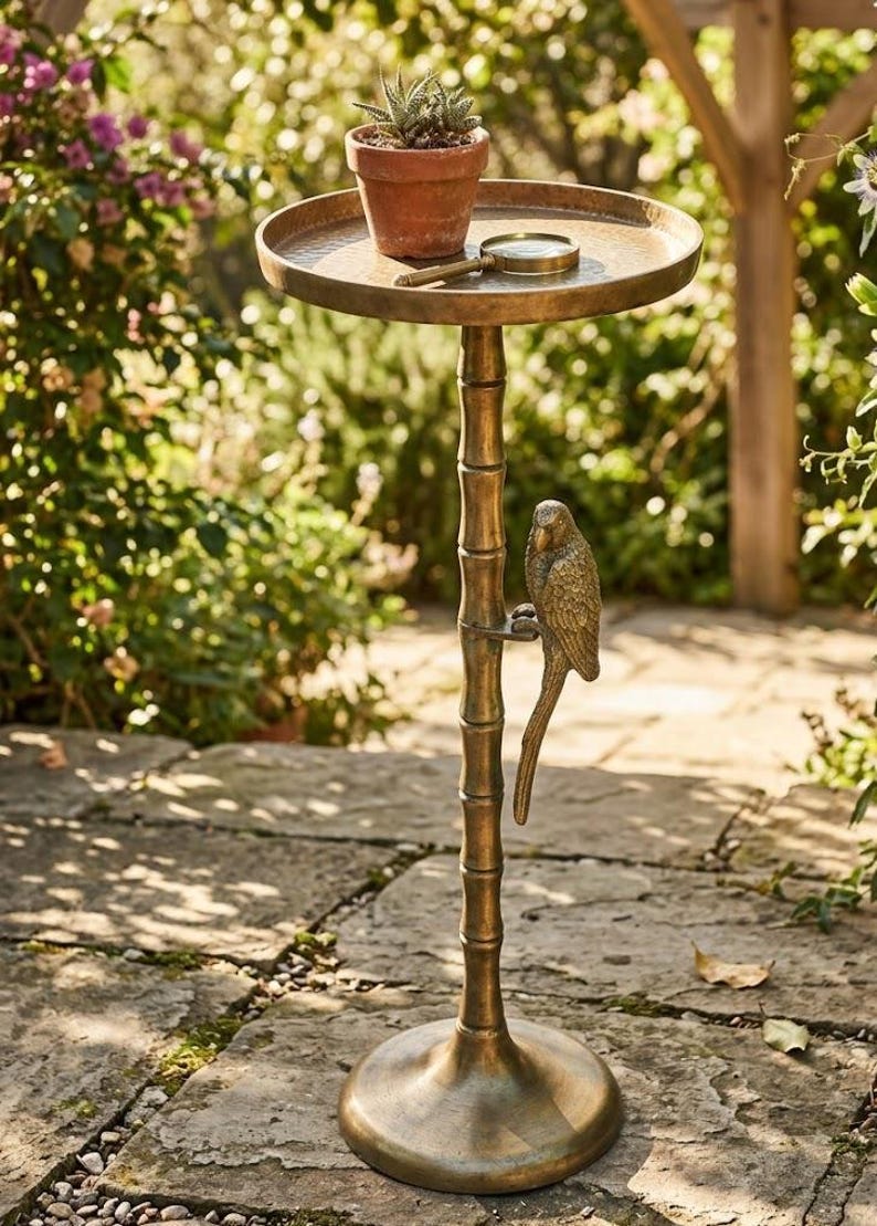 May include: A bronze-colored plant stand with a round top, a bamboo-style stem, and a parrot figurine. A small potted plant and a magnifying glass rest on the top. The stand is outdoors on a stone patio.