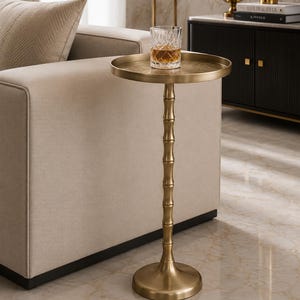 May include: A tall, round side table with a metallic gold finish and a textured, bamboo-inspired central pedestal base. The table features a flat, circular top surface and a wide, stable circular base, supporting a clear, faceted glass tumbler.