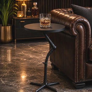 May include: A black side table with a round top and a base shaped like a bird's leg and foot. The table is positioned next to a brown leather sofa and holds a clear glass tumbler filled with an amber liquid and ice cubes.