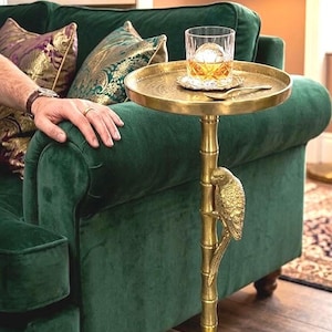 Könnte beinhalten: Ein goldfarbener Beistelltisch mit runder Tablettplatte, getragen von einer Bambusstange mit Papageienakzent. Der Tisch trägt ein Glas bernsteinfarbener Flüssigkeit mit einer großen Eiskugel. Der Tisch steht neben einem grünen Samtsofa.