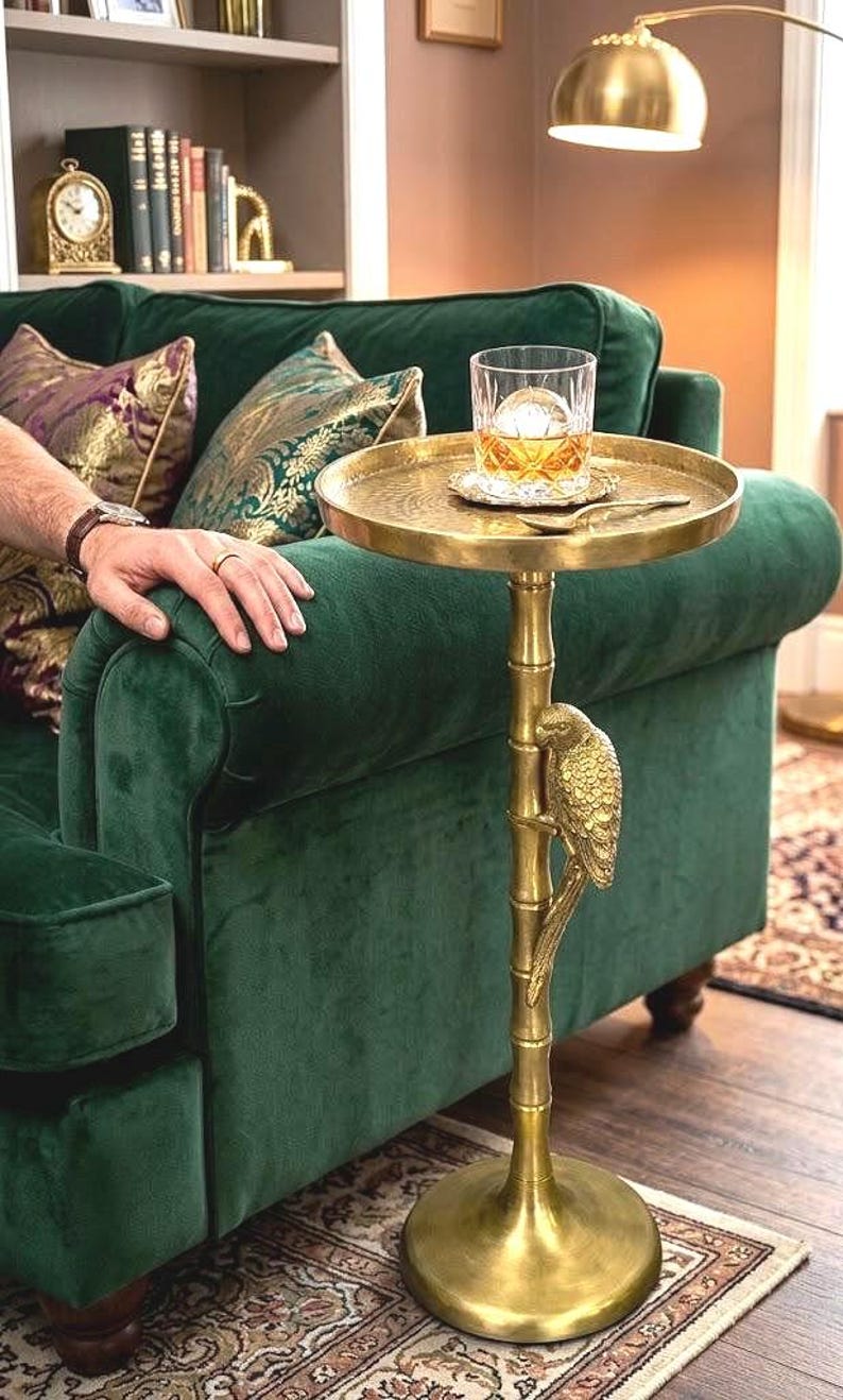 May include: A gold-colored side table with a round tray top, supported by a bamboo-style pole with a parrot accent. The table holds a glass of amber liquid with a large ice sphere. The table is next to a green velvet sofa.