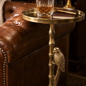 May include: A gold-colored side table with a round tray top. A cut-crystal glass with amber liquid sits on the tray, along with a pen and a small notebook. The table's stem is bamboo-shaped, with a parrot sculpture.