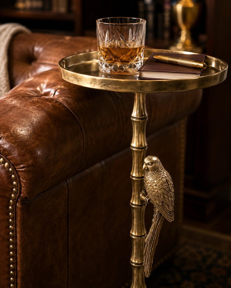 May include: A gold-colored side table with a round tray top. A cut-crystal glass with amber liquid sits on the tray, along with a pen and a small notebook. The table's stem is bamboo-shaped, with a parrot sculpture.