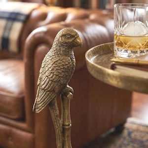 May include: A detailed brass parrot sculpture perched on a bamboo-style stand. The bird has intricate feather detailing and a curved beak. A round, gold-toned tray holds a glass of whiskey with a large ice sphere. The background features a leather sofa.