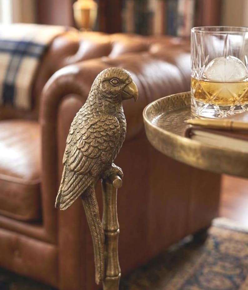 May include: A detailed brass parrot sculpture perched on a bamboo-style stand. The bird has intricate feather detailing and a curved beak. A round, gold-toned tray holds a glass of whiskey with a large ice sphere. The background features a leather sofa.