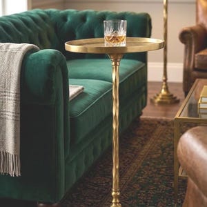 Pode incluir: Uma mesa lateral dourada com um tampo redondo e uma haste torcida. Um copo de líquido âmbar está sobre a mesa. A mesa está ao lado de um sofá de veludo verde e um tapete estampado.