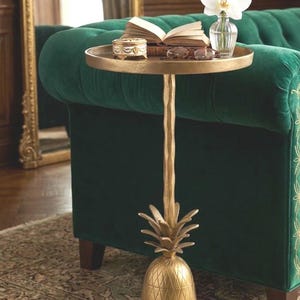 May include: A gold-colored side table with a pineapple base and a round top. The table holds an open book, glasses, a small decorative box, and a vase with a white orchid. The table is in front of a green velvet sofa.