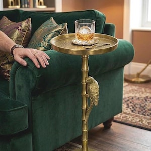 May include: A gold-colored side table with a round top, supported by a bamboo-style stem and a decorative parrot. A glass of whiskey with a large ice sphere sits on the table. The table is next to a green velvet sofa.