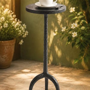 Puede incluir: Pequeña mesa auxiliar de metal negro con tapa redonda y base de tres patas. Una taza de café blanca y un platillo con una cuchara descansan sobre la mesa. La mesa está al aire libre, cerca de plantas en macetas.