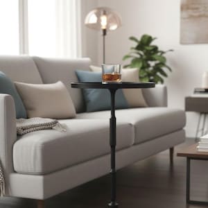 Może przedstawiać: Czarny stolik pomocniczy z okrągłym blatem, na którym stoi szklanka bursztynowego płynu z lodem. Stolik stoi przed szarą sofą z poduszkami. Stolik ma około 76 cm wysokości.