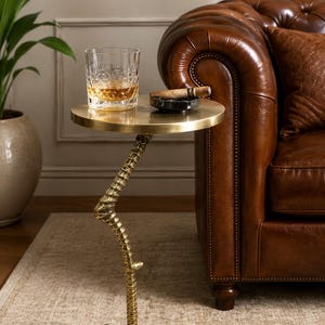 May include: A round brass side table featuring a base shaped like a bird's leg with talons. The table top holds a clear crystal glass filled with an amber liquid and a black ashtray containing a brown cigar, showcasing a metallic finish.