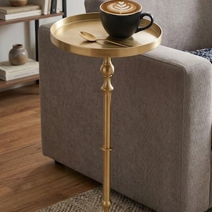 Puede incluir: Una mesa auxiliar dorada con una parte superior redonda y un solo pie. Una taza de café negra con arte latte y una cuchara descansan sobre la mesa. La mesa está al lado de un sofá gris y una alfombra estampada.