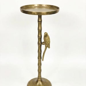 May include: A gold-colored side table with a round top and a bamboo-style stem. A detailed parrot figure is perched on the stem. The table has a circular base and is made of metal.