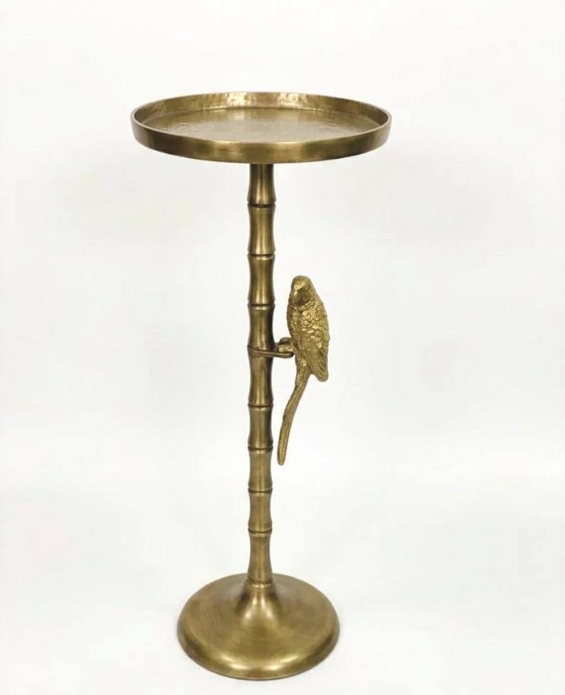 May include: A gold-colored side table with a round top and a bamboo-style stem. A detailed parrot figure is perched on the stem. The table has a circular base and is made of metal.