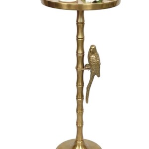 May include: A gold-colored side table with a round top and a bamboo-style stem. A decorative parrot is perched on the stem. A glass with a drink and a white flower are on the table's surface.