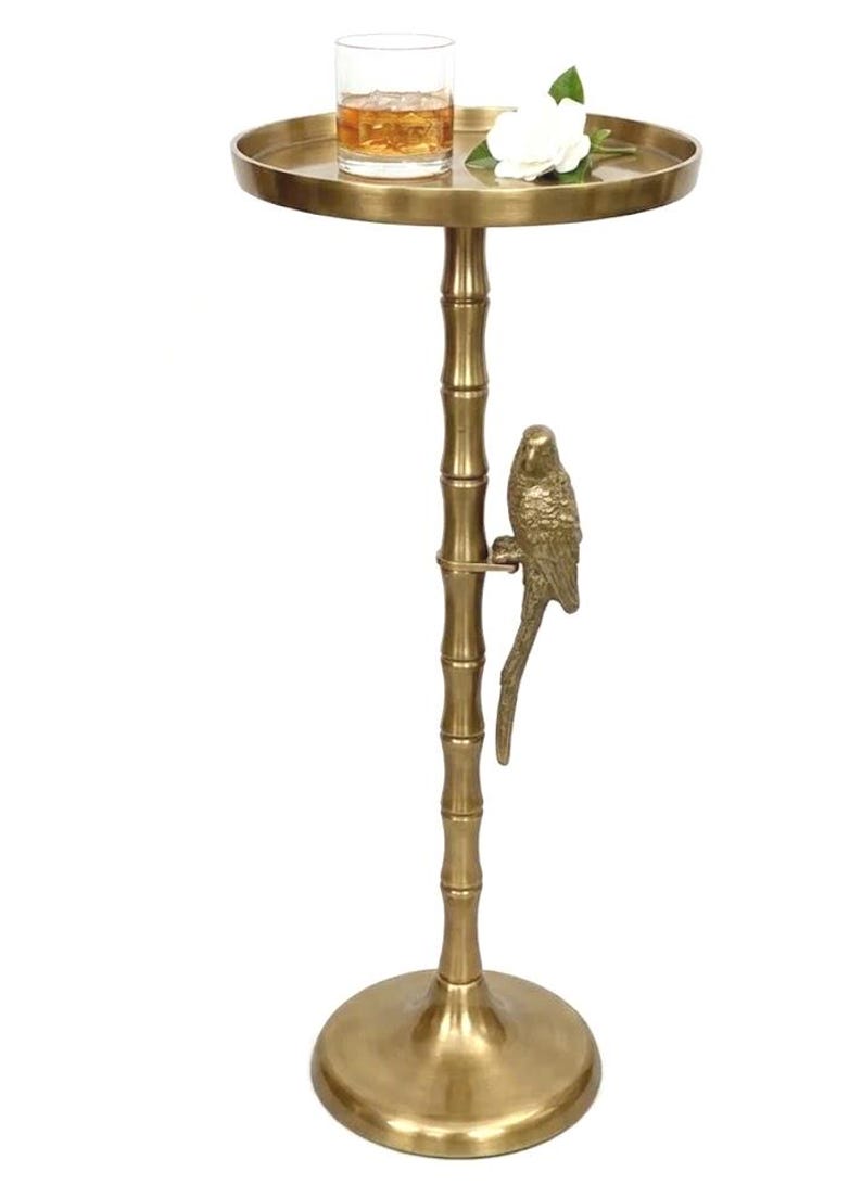 May include: A gold-colored side table with a round top and a bamboo-style stem. A decorative parrot is perched on the stem. A glass with a drink and a white flower are on the table's surface.