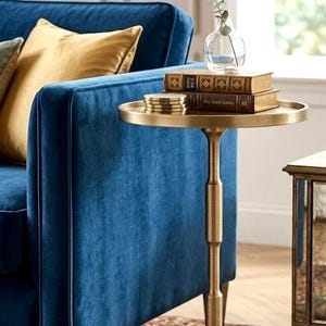 Puede incluir: Una mesa auxiliar dorada con una tapa redonda que sostiene libros y posavasos. La mesa tiene un tallo acanalado y una base circular. Un sofá de terciopelo azul y una alfombra estampada están en el fondo. Un jarrón de cristal con vegetación está sobre la mesa.