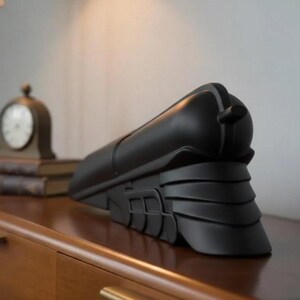 May include: A black, streamlined object resembling a train engine, resting on a wooden surface. The object has a rounded top, a series of layered panels at the base, and a small, cylindrical protrusion at the rear. A clock and books are in the background.