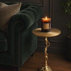May include: A lit candle in a glass jar sits on a small, gold side table with a unique, sculptural base. The table is next to a dark green velvet sofa with a patterned pillow. The candle label reads "SANTAL 26".