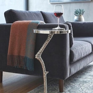 May include: A silver side table with a bird claw base and a round top. A glass of red wine sits on the table next to a dark grey sofa with a fringed throw. The table is a decorative accent piece, ideal for a living room.