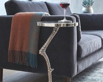 Può includere: Un tavolino d'argento con base a zampa di uccello e piano rotondo. Un bicchiere di vino rosso è sul tavolo accanto a un divano grigio scuro con una coperta con frange. Un elemento decorativo.