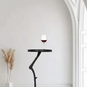 May include: A black accent table with a unique, bird-like leg design. The round tabletop holds a glass of red wine. The table is in a bright, minimalist room with a vase of dried plants.