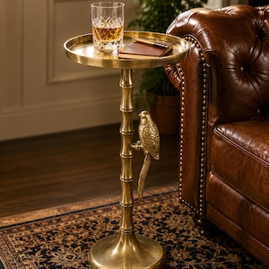 May include: A gold-colored side table with a round top and a parrot-shaped accent. A glass of amber liquid and a small notebook rest on the table. The table has a bamboo-style stem and a round base.