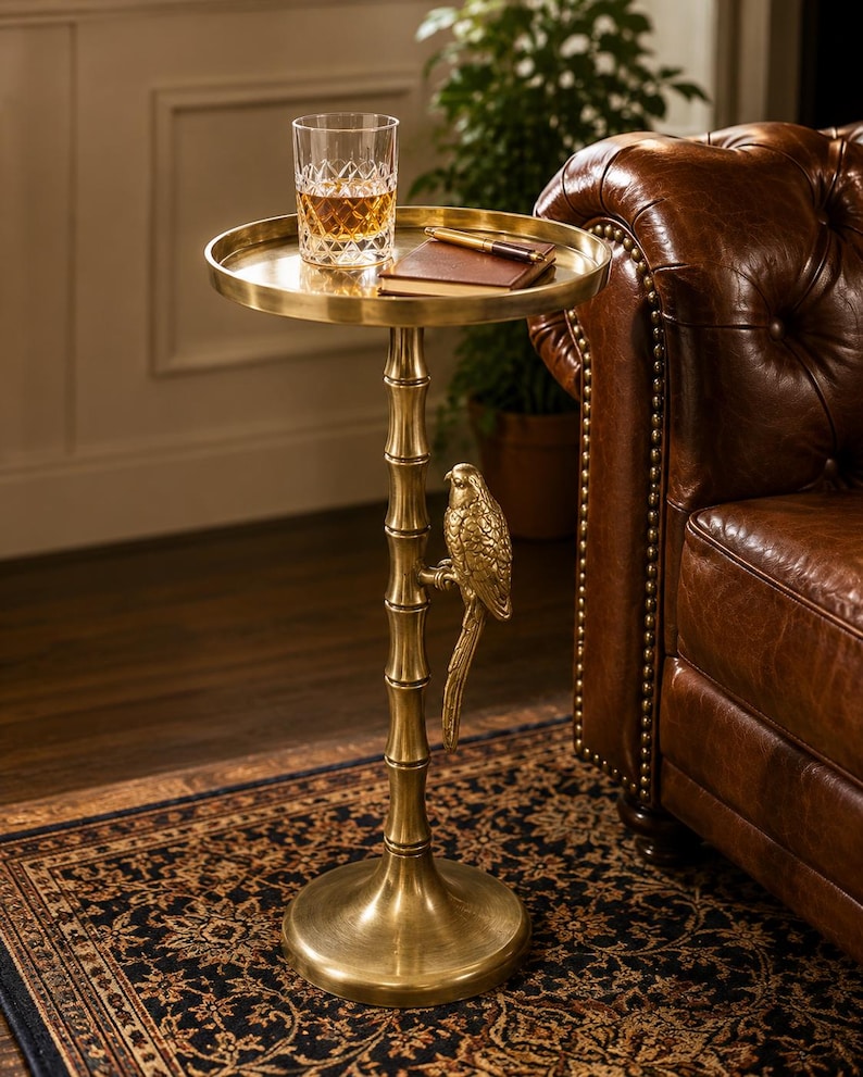 May include: A gold-colored side table with a round top and a parrot-shaped accent. A glass of amber liquid and a small notebook rest on the table. The table has a bamboo-style stem and a round base.