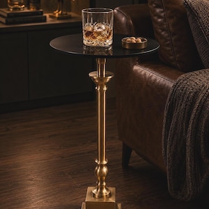 Puede incluir: Una mesa auxiliar redonda de color negro con una base de pedestal alta y delgada de color latón con una base cuadrada. La mesa sostiene un vaso de cristal con un líquido ámbar y una esfera de hielo, junto a un pequeño cuenco de latón con frutos secos.