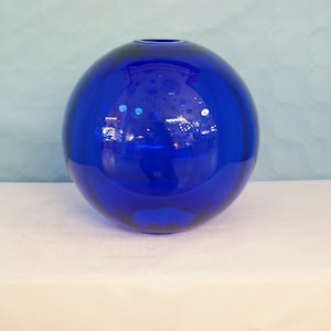 以下が含まれることがあります： 球形の濃い青色のガラスの花瓶。花瓶は白い表面の上に置かれています。
