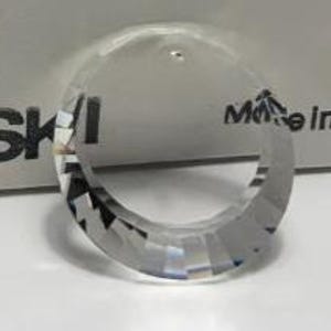 Puede incluir: Un anillo de cristal transparente y facetado, que refleja la luz. El anillo es circular con un diseño geométrico. El fondo presenta una caja blanca con la palabra "SWAROVSKI" y "Made in Austria" impresas.