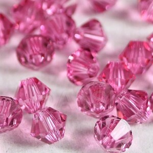 Könnte beinhalten: Nahaufnahme einer Sammlung facettierter, rosa Swarovski-Kristalle. Die Kristalle sind auf einer weißen Oberfläche verstreut, mit dem Wort "SWAROVSKI" in Grau am unteren Rand. Die Kristalle haben mehrere Facetten, die das Licht reflektieren.