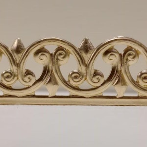 May include: A gold-coloured decorative trim with a repeating, ornate pattern. The design features connected swirls and pointed elements, creating a detailed border. The trim is likely used for embellishment.