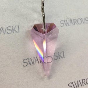 May include: A light pink crystal pendant with a silver-coloured metal bail. The crystal is a long, pointed shape with multiple facets that catch the light, creating a rainbow effect. The background has the word "SWAROVSKI" printed on it.