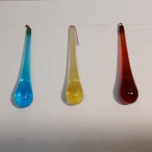 May include: Five colorful glass teardrop ornaments in green, blue, yellow, red, and pink. Each ornament has a small gold-colored loop at the top for hanging. The teardrops are smooth and translucent.