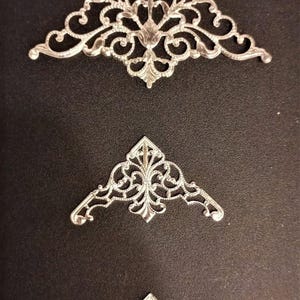 Può includere: Tre decorazioni angolari argentate e ornate di varie dimensioni sono esposte su uno sfondo scuro. Ogni pezzo presenta un design simmetrico dettagliato con motivi a spirale e un motivo floreale centrale, adatto per progetti di artigianato.